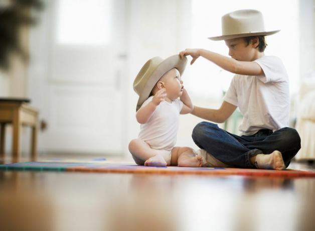 selektiv fokus bild av två bröder, en är nio en är åtta månader paret sitter med korsbenen på golvet äldre pojke sätter matchande hatt på barnets huvud