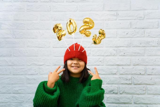 glad asiatisk flicka i en grön tröja närbild med gyllene ballonger med siffror 2023 i ett hattkopiautrymme, vit tegelväggbakgrund, flicka tittar på kameran, ler