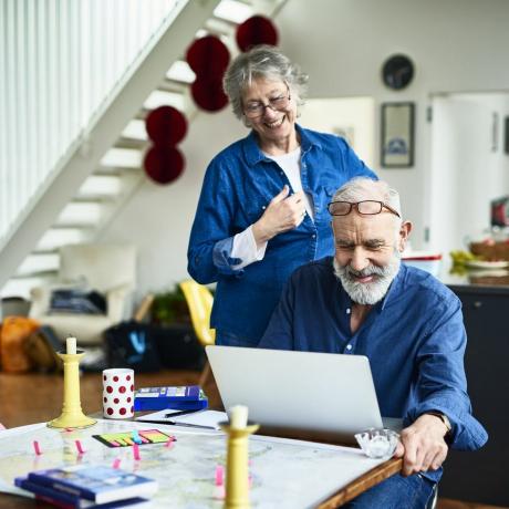 äldre kvinna ler och tittar över makens axel när han forskar och läser recensioner online om övernattningsställen och platser att besöka, klisterlappar och guideböcker på bordet, bokning av semester uppkopplad