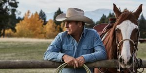 kevin costner i yellowstone bredvid en häst lutad på ett staket med rep i händerna iklädd en blekt blå jeansskjorta och beige cowboyhatt