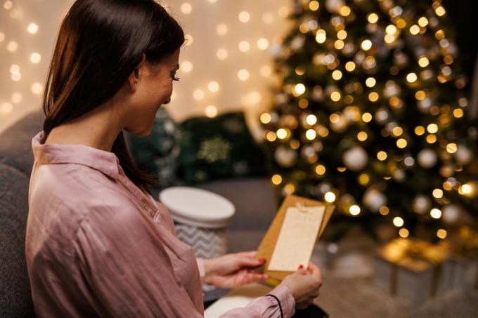 glad ung kvinna sitter i soffan, vid den glittrande julgranen, öppnar och läser ett julkort hon fick med sin present