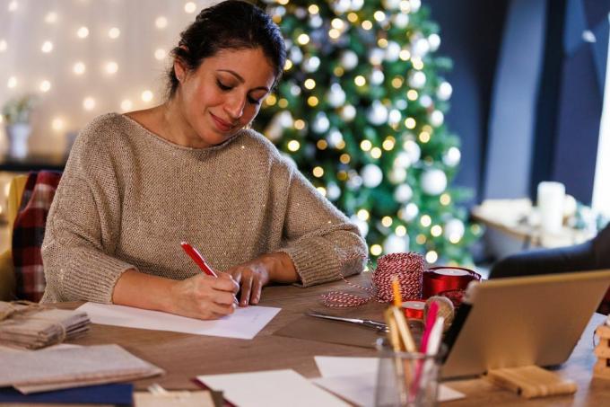porträtt av en glad mitten av vuxen kvinna som sitter vid bordet, framför en glittrande julgran, ler medan du skriver julkort och brev till nära och kära