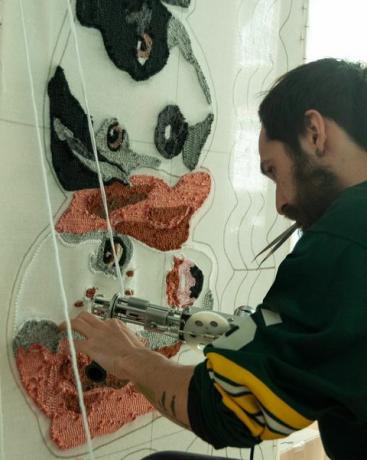 30-årig spansk latino-skäggig man klädd i en rugbytröja, i sin verkstad och arbetar på handgjorda mattor gjorda med tuftpistol och akrylfibrer, med en hundmattadesign
