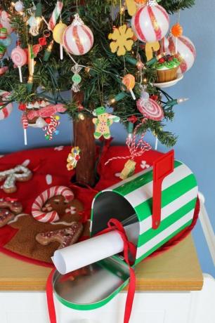 liten vit och grön randig brevlåda med rullat papper bundet med ett rött band i det, sitter under ett litet julgran, på en röd julgran kjol