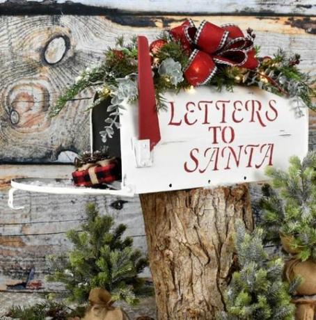 vit brevlåda med "brev till santa" med röda bokstäver skrivna på den, sitter på en stock, dörren till den är öppen och en present sprider ut ur den är brevlådan toppad med julgrönt och ett rött rosett