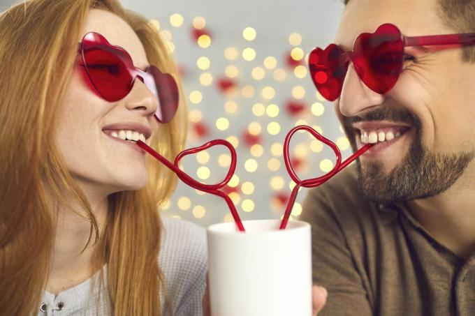 närbild porträtt glad ung man och kvinna förälskad i rosa solglasögon smuttar på drink från en kopp genom hjärtformade sugrör, njuter av söta roliga ögonblick på en rolig dejt på Sankt Alla hjärtans dag