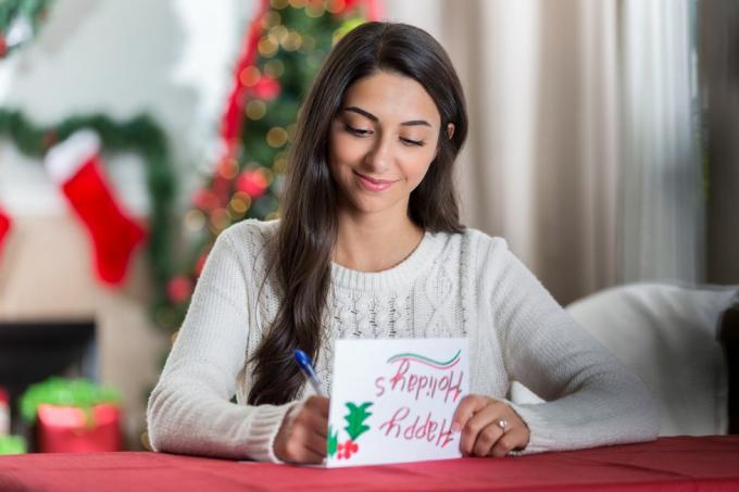 en leende ung kvinna sitter vid ett bord i sitt dekorerade vardagsrum och tittar koncentrerat ner när hon skriver ett brev till en vän till jul