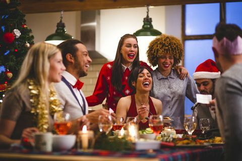 en grupp vänner som skrattar när de sitter runt ett bord och äter och dricker på julen en man läser ett skämt från en julknäcka festhattar och jultröjor bärs